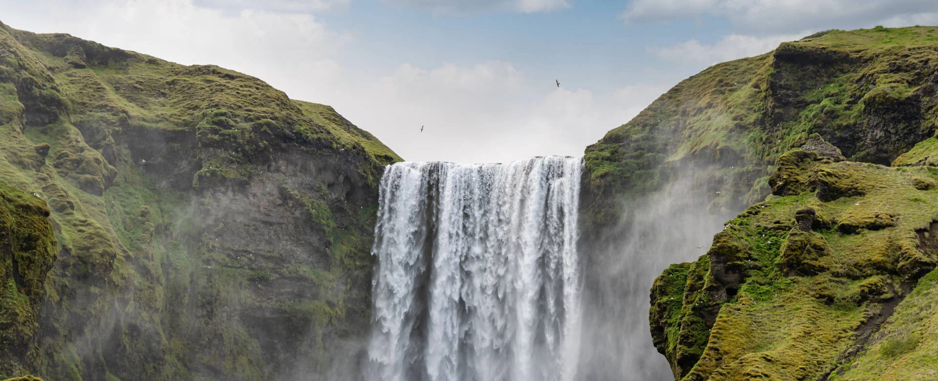 Ein beeindruckender Wasserfall stürzt zwischen zwei grasbewachsenen Klippen herab, umgeben von einem bewölkten Himmel und Grün.
