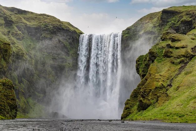 Ein beeindruckender Wasserfall stürzt zwischen zwei grasbewachsenen Klippen herab, umgeben von einem bewölkten Himmel und Grün.
